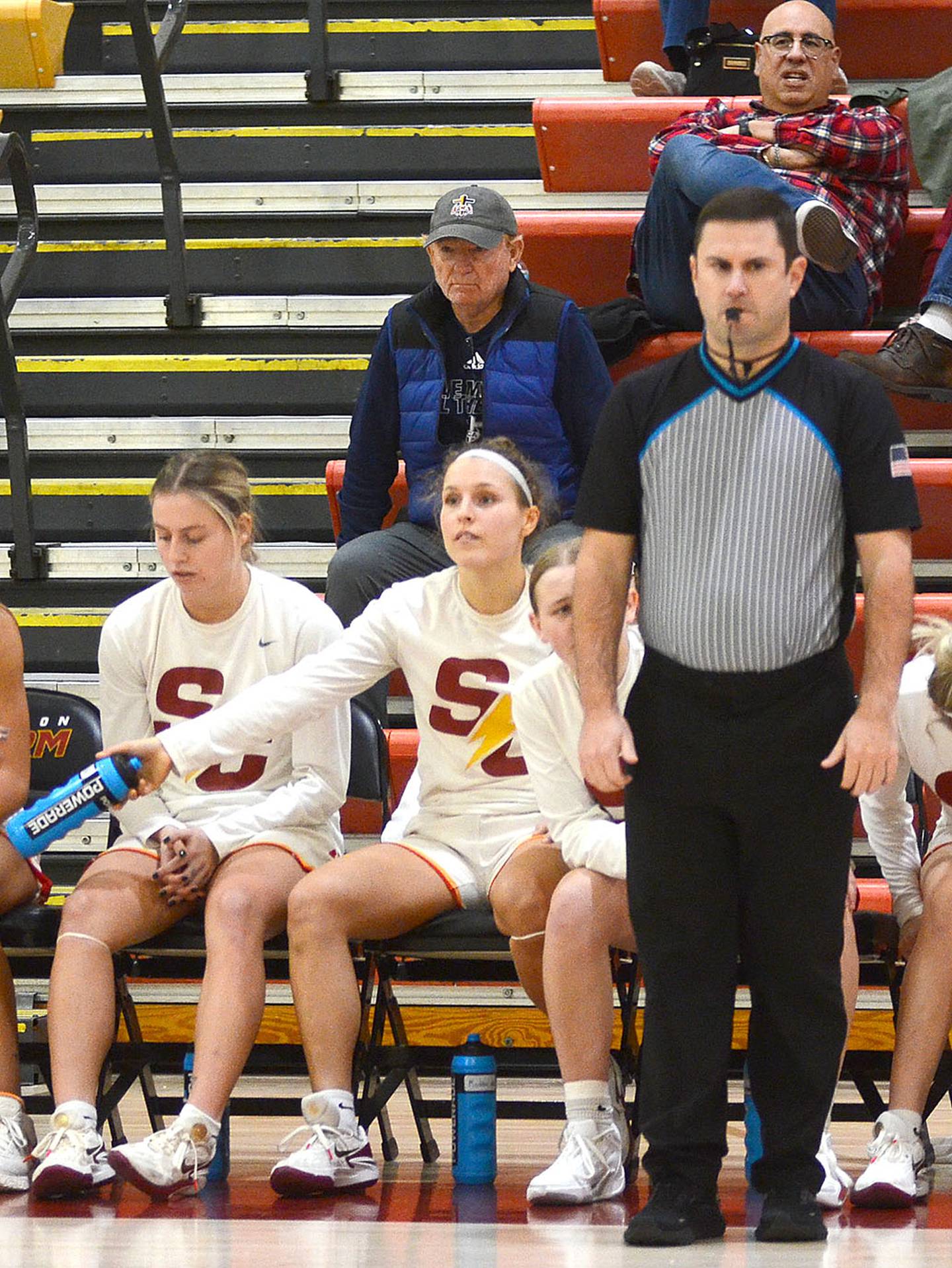 Gerry Miller, Nodaway Valley High School principal, is shown officiating Wednesday's Simpson game against Coe. Maddax DeVault, Simpson sophomore from Greenfield, is seated on the bench near Miller.